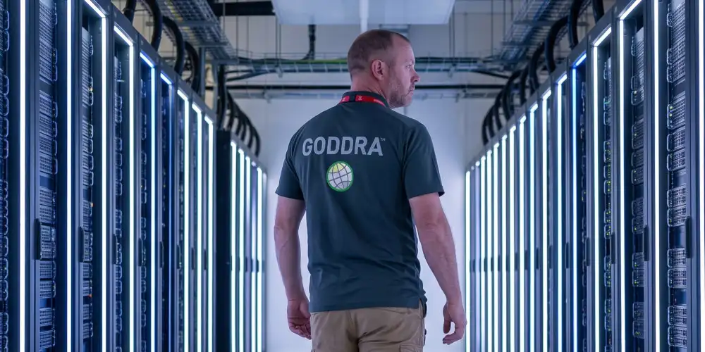 A GODDRA cybersecurity officer wearing a branded t-shirt viewed from behind as he walks down a server room corridor.