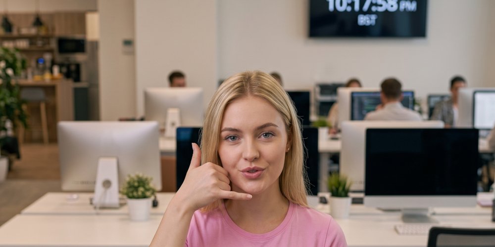 A GODDRA team member in a modern open-plan office, making the 'call me' gesture and smiling, with a global network logo on her pink t-shirt.