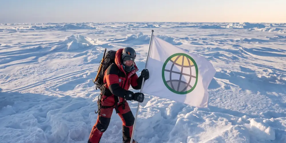 An arctic explorer clad in extreme-weather survival gear planting a green, yellow, and white GODDRA branded flag firmly into the snow at a vast, icy expanse at the North Pole.