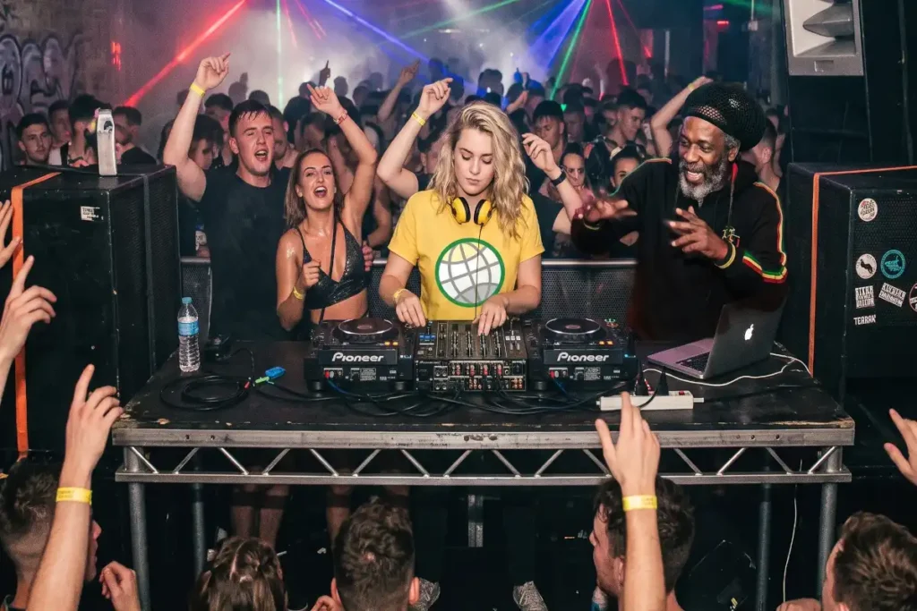 DJ wearing a yellow GODDRA branded t-shirt performing closely in front of ravers in an intimate nightclub MC is readying for bars.